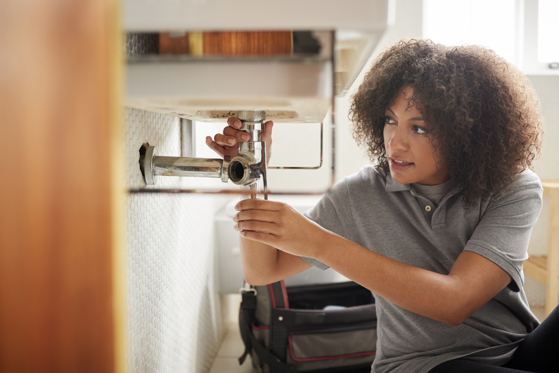 Young black female plumber sitting on the floor fixing a bathroom sink, seen from doorway