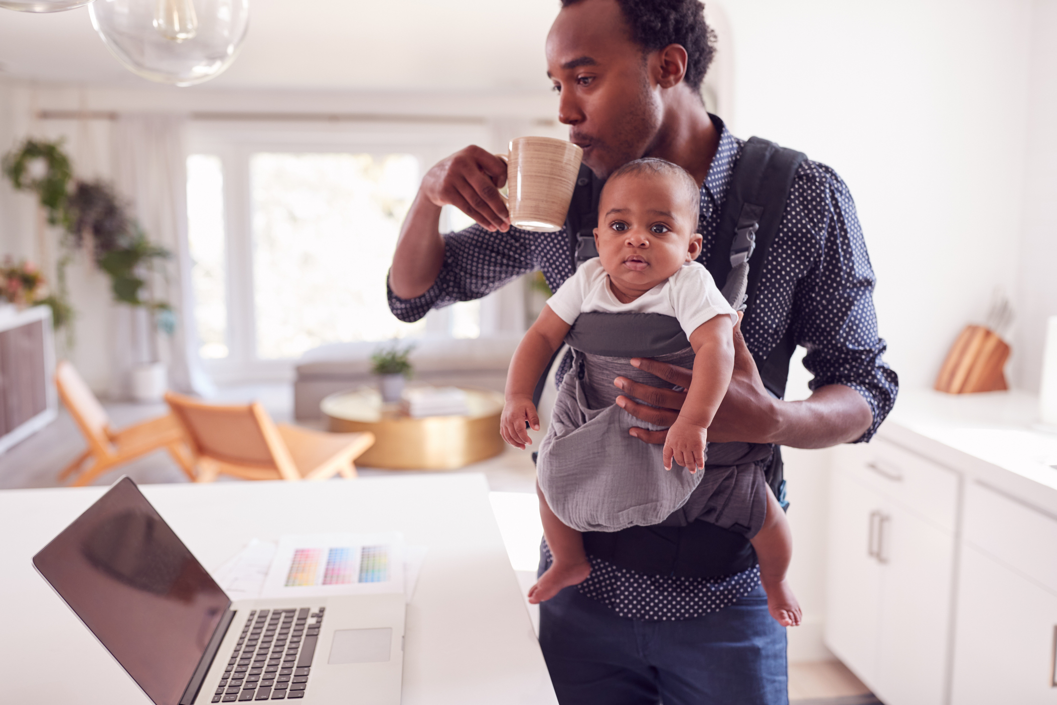 Father With Baby Daughter In Sling Multi-tasking Working From Home On Laptop