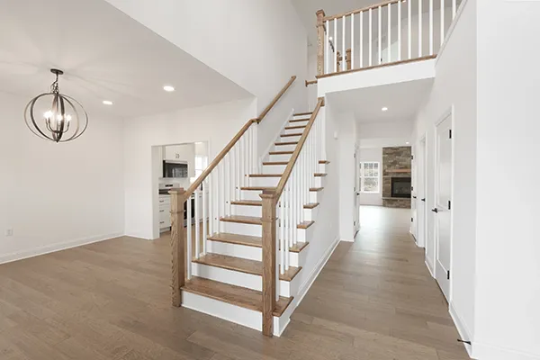 Bayberry foyer with open staircase with oak treads, white risers, wood handrails, and vinyl plank flooring. View into kitchen and family room area with fireplace. 